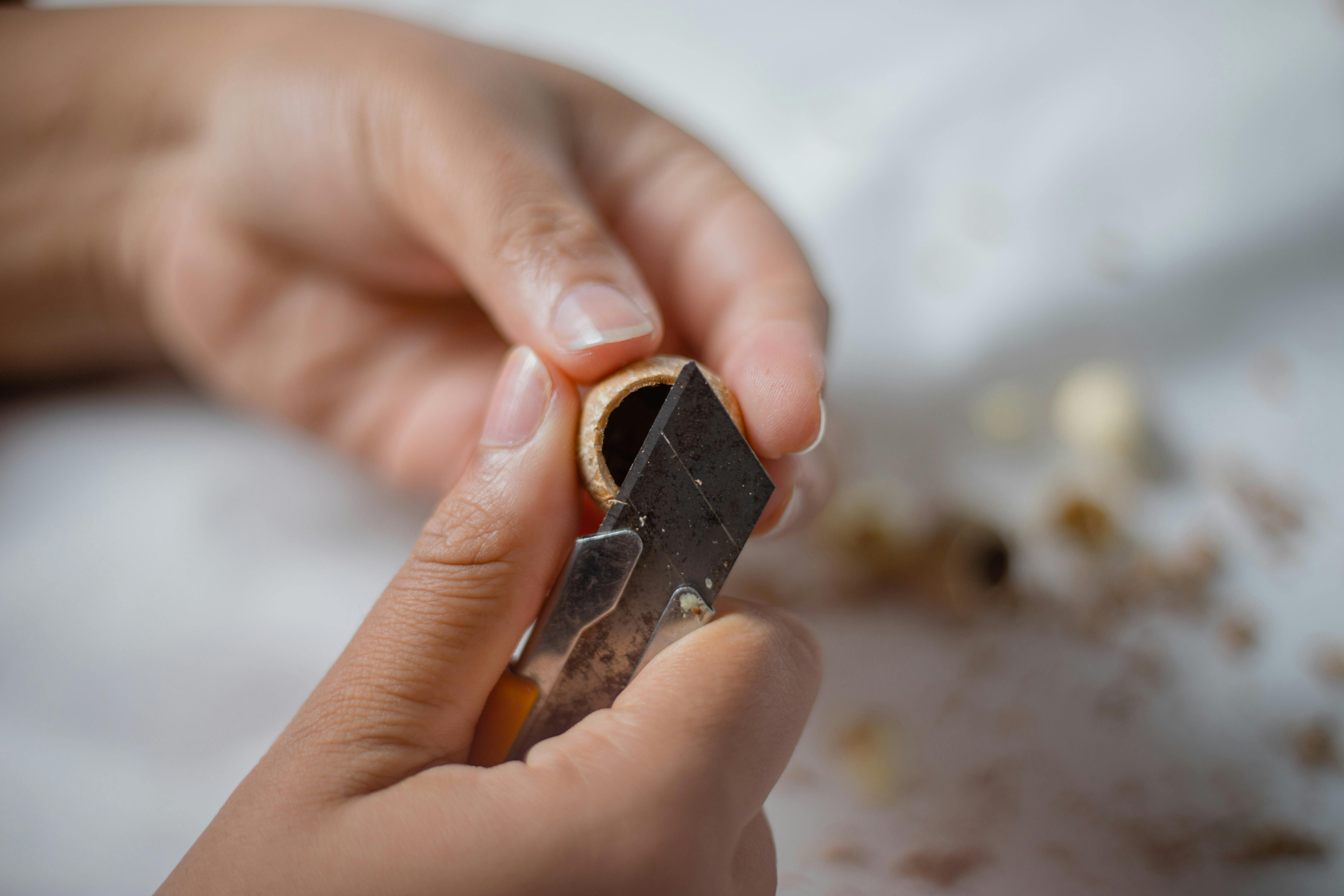 Crop anonymous master cutting wooden shell with sharp stationery knife while working at table on blurred background in light workshop